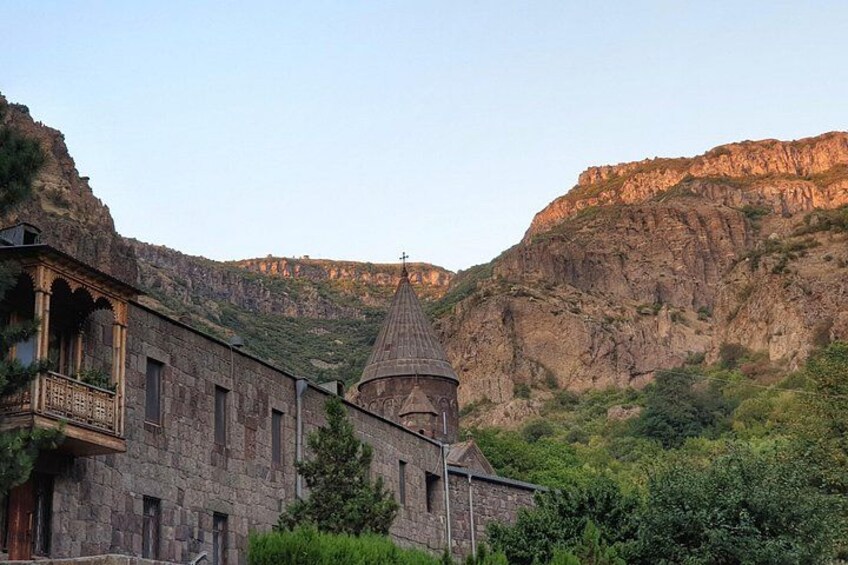 A stone building with a bell tower, surrounded by greenery and mountains. 4
