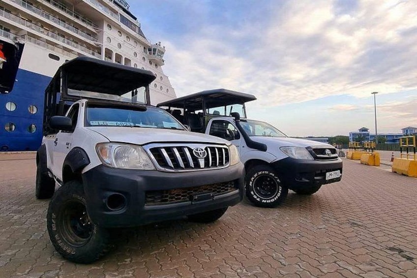Two off-road vehicles parked on a paved surface with a cruise ship in the background. 1