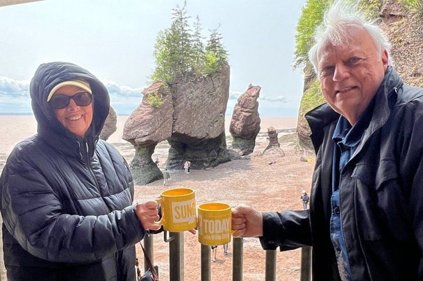Two people holding yellow mugs with the word 'TODAY' on them, standing in front of a rocky landscape with large boulders and a body of water in the background. 2