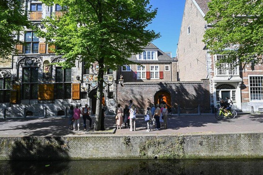 Een historisch gebouw met een grote boog, een boom op de voorgrond en mensen die op het trottoir staan. 1