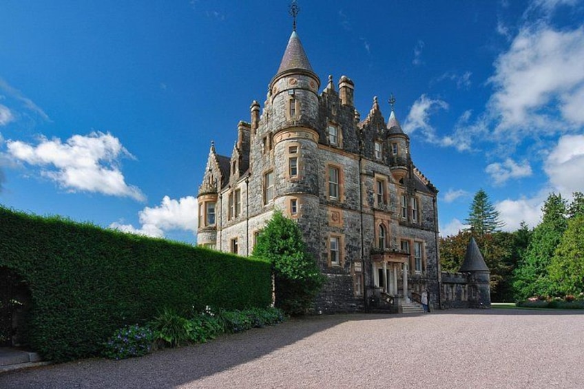 A large, historic stone castle with multiple towers and turrets, surrounded by well-maintained hedges and a gravel driveway. 1