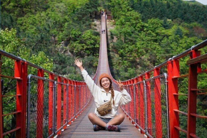 Una persona sentada sobre un puente colgante rojo, con follaje verde al fondo. 1