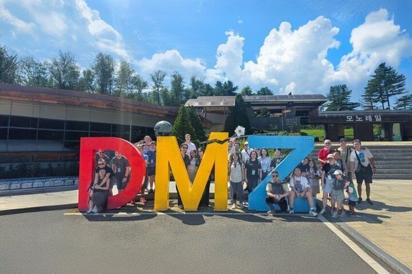 Un grupo de personas posando con letras grandes y coloridas que deletrean 'DMZ' en un estacionamiento. 4
