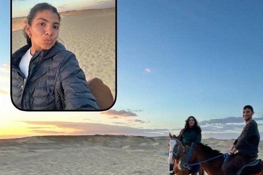 En person i en blå jakke tager et selfie, og to personer rider på heste i et ørkenlandskab med klitter og en klar himmel. 2