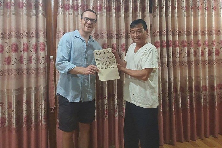 Two men holding a sign with text, standing in front of a curtain with a floral pattern. 3