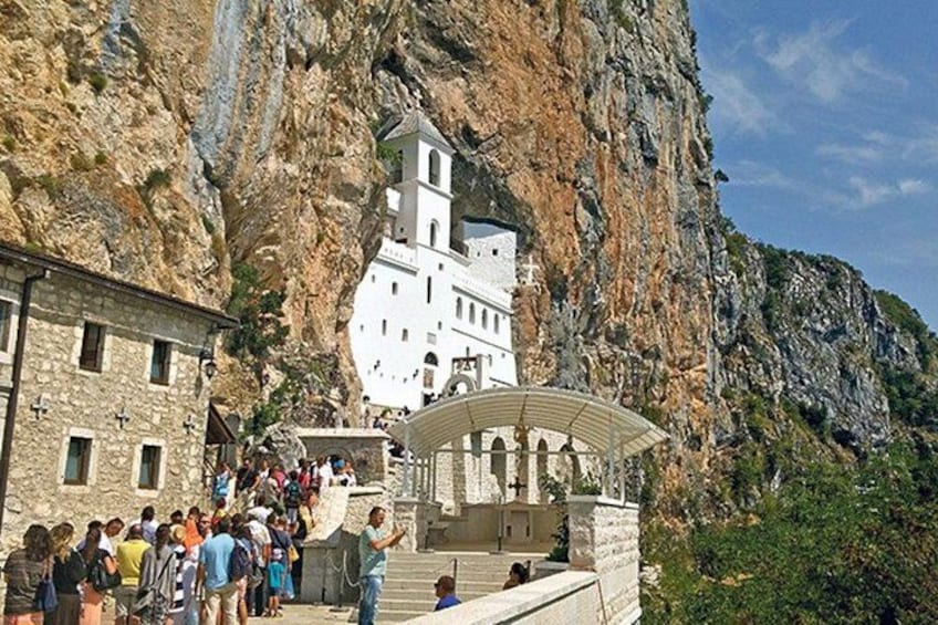 A white church built into a cliffside, a stone building in the foreground, and a group of people gathered at the base. 5