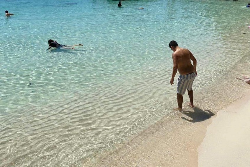 A beach with clear water, people swimming, and a person standing on the sand. 3