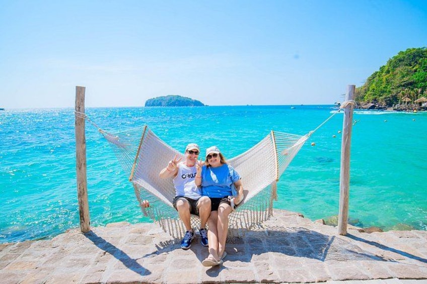 Two people sitting on a hammock by a turquoise sea with a rocky shore and a small island in the distance. 1