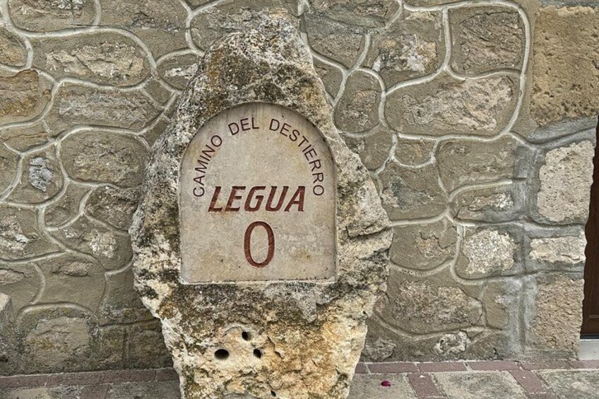 A stone monument with the inscription 'CAMINO DEL DESIERRO LEGUA 0' against a stone wall background. 3