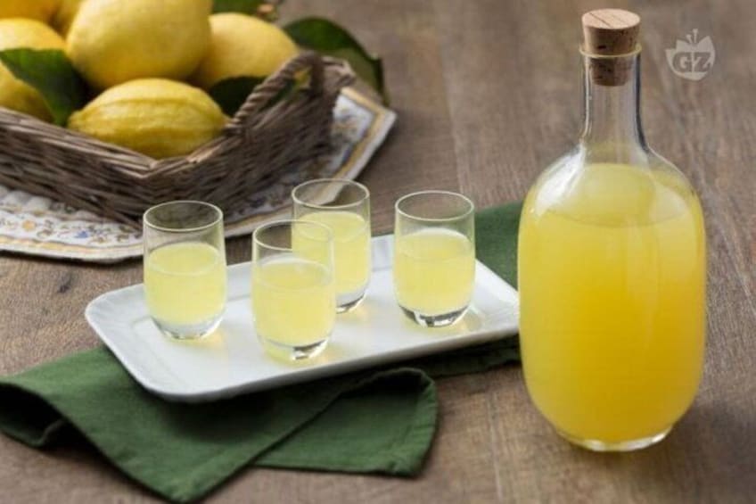 A bottle of yellow liquid, possibly lemonade, on a wooden table with a green cloth, accompanied by a plate with shot glasses and lemons in the background. 2
