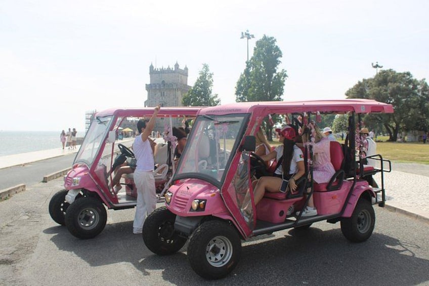 Pink golf carts with passengers on a paved path near a body of water and a castle-like structure in the background. 3