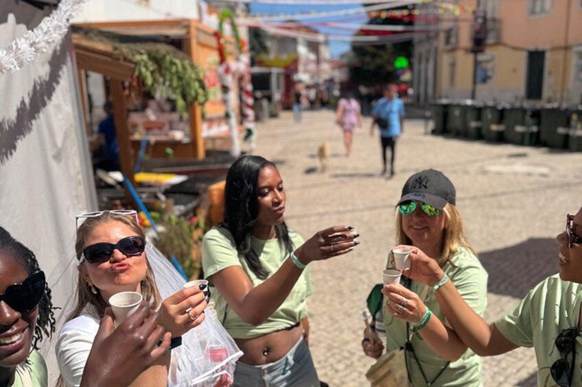 A group of people toasting with drinks in an outdoor setting with market stalls and a cobblestone street. 2