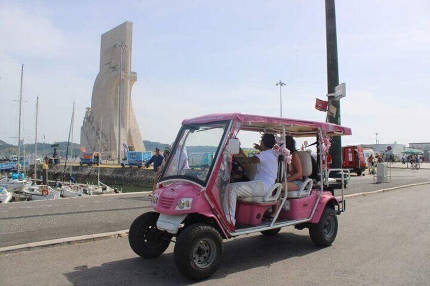 Ein pinkfarbenes, dreirädriges Fahrzeug auf einer Straße mit einem monumentalen, segelartigen Bauwerk im Hintergrund. 1