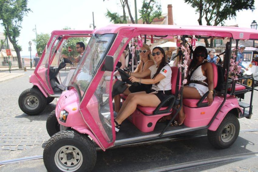 A pink trolley with passengers, a cobblestone street, and trees in the background. 1