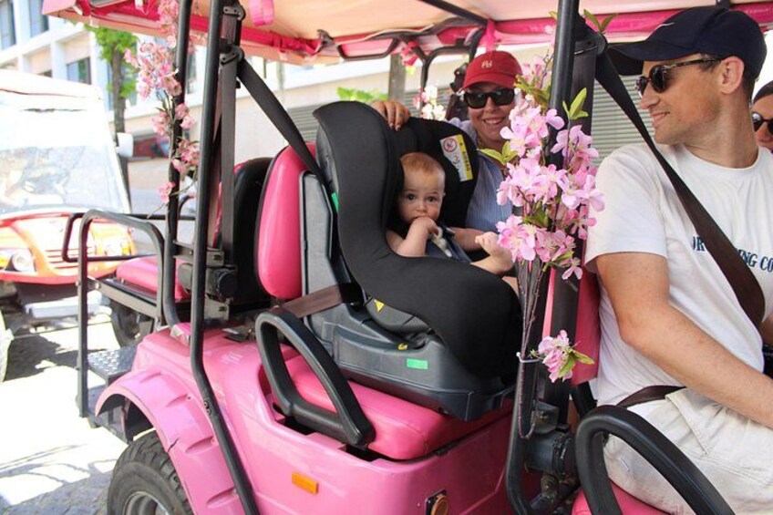 A pink golf cart with a child seated in the back, a man driving, and a woman standing beside. 2