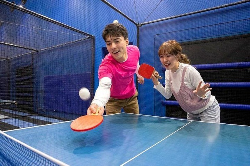 Two people playing table tennis in an indoor court with blue walls and a net. 2