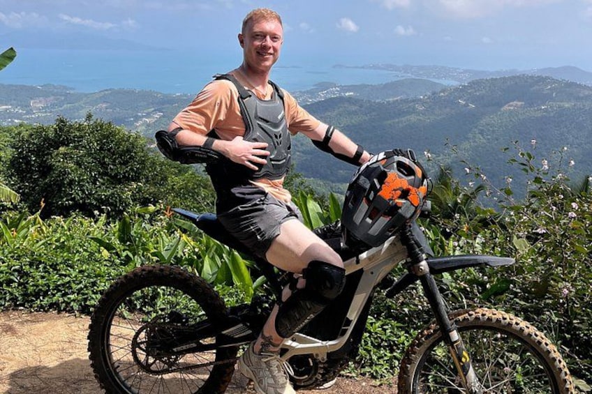 En person sidder på en mountainbike med hjelm og udstyr, omgivet af grøn vegetation og et smukt udsigt over bakker og vand. 3