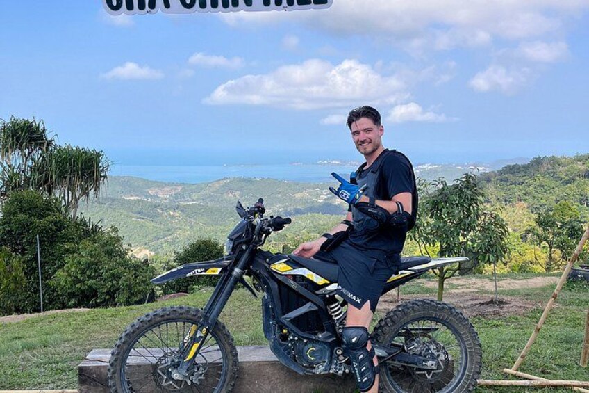 En person sidder på en dirtbike med udsigt til bakker og et vandareal i baggrunden. 5