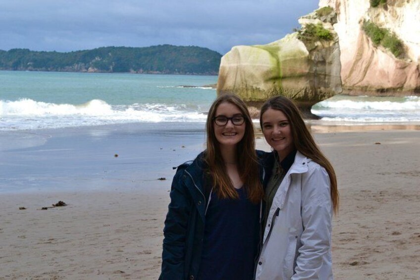 Dos personas de pie en una playa con una gran formación rocosa y el océano al fondo. 3