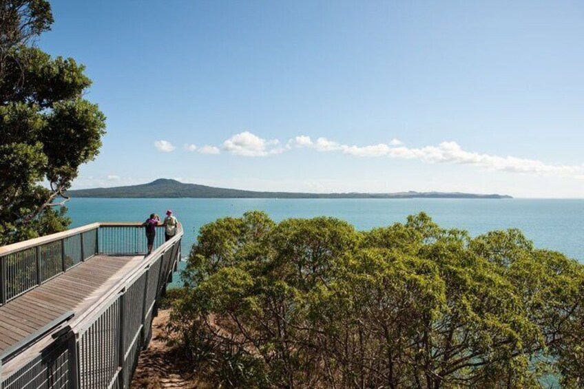 A wooden boardwalk with people walking, overlooking a calm sea and distant landmass. 4