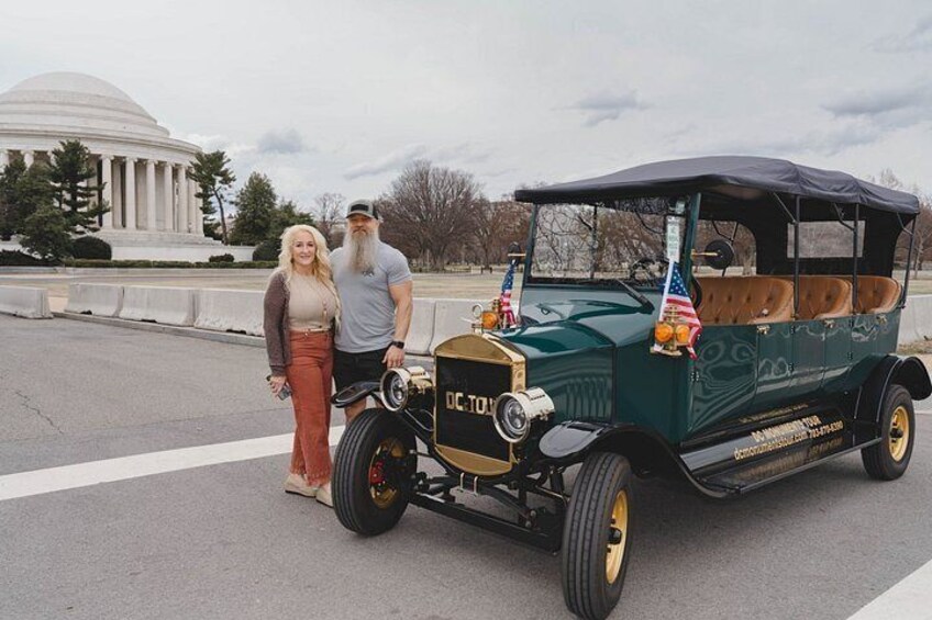 A vintage car with a wooden interior, parked on a road with a monument in the background. 2