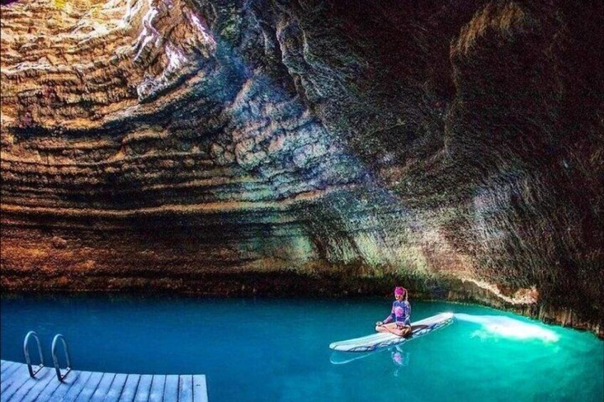 A person in a swimsuit sitting on a floating platform in a clear blue pool surrounded by rocky cave walls. 1