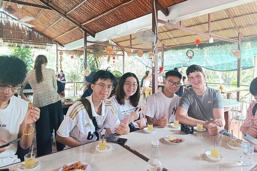 A group of people dining at an outdoor restaurant with a thatched roof and tables set with plates, glasses, and cutlery. 3