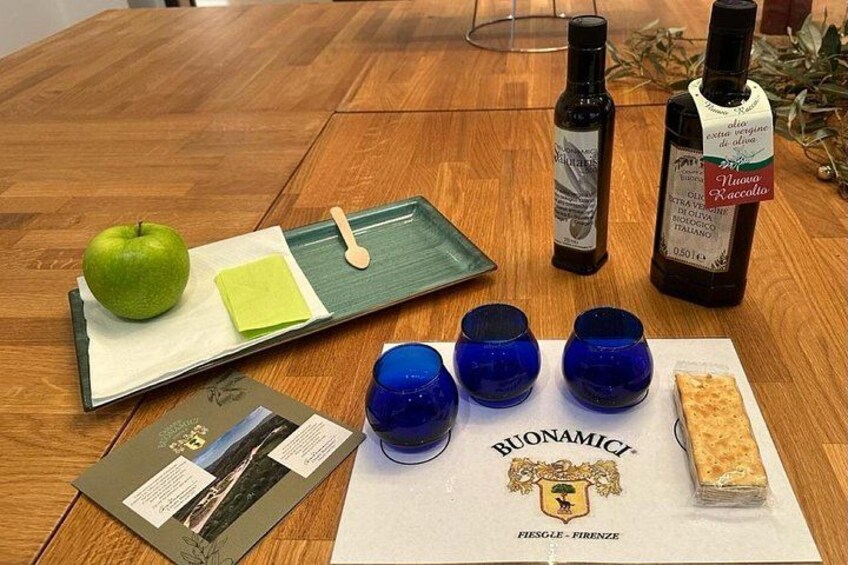 Een houten tafel met een groene dienblad, een groene appel, een fles olijfolie, blauwe glazen, een boekje en een rechthoekig stuk kaas. 1