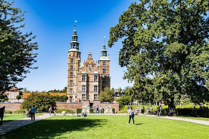 En historisk byggnad med två torn, omgiven av en välskött park där människor promenerar och sitter på bänkar. 2