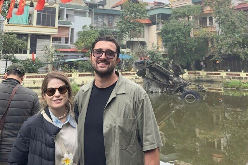 A man and a woman standing in front of a damaged vehicle in a water body, with buildings in the background. 3