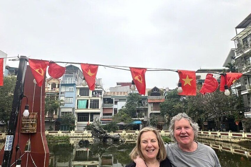 Two people in front of a traditional boat with red flags and a water body. 4