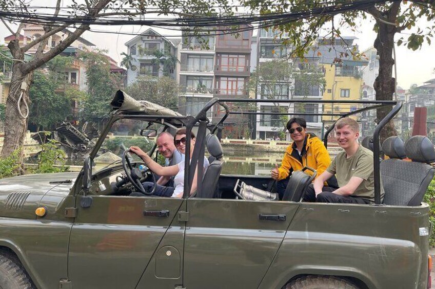 Four people in a green jeep with an open top, parked in an urban area with buildings and trees in the background. 5