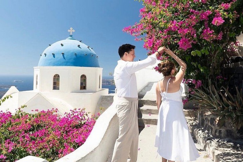 Un couple danse sur le toit d’un bâtiment blanchi à la chaux, surmonté d’un dôme bleu, et entouré de fleurs roses et d’un ciel d’un bleu limpide. 1