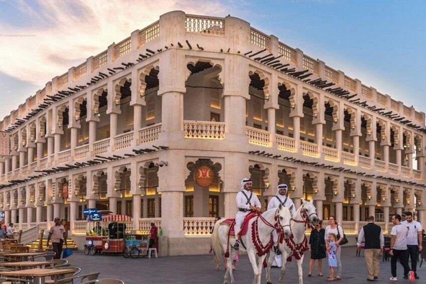 Un grand bâtiment imposant, richement décoré, doté de nombreux balcons et de colonnes, des chevaux avec des cavaliers vêtus de costumes traditionnels, et des gens qui se promènent devant. 1