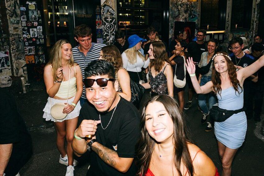 A group of people at a social gathering, possibly a party or event, with a wall decorated with various posters and pictures. 3
