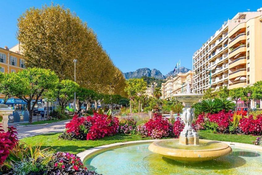 Una fuente rodeada de flores y árboles vibrantes, con edificios y montañas al fondo. 2