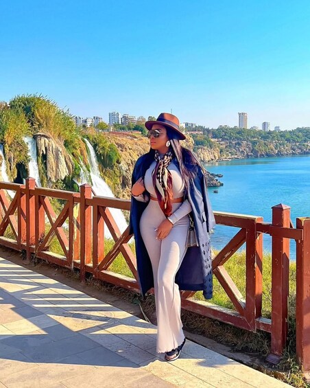 A person sitting on a wooden railing by a coastal area with a waterfall and buildings in the background. 2