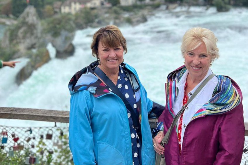 Twee personen staan bij een hek met een rivier en een rotsachtig landschap op de achtergrond. 1