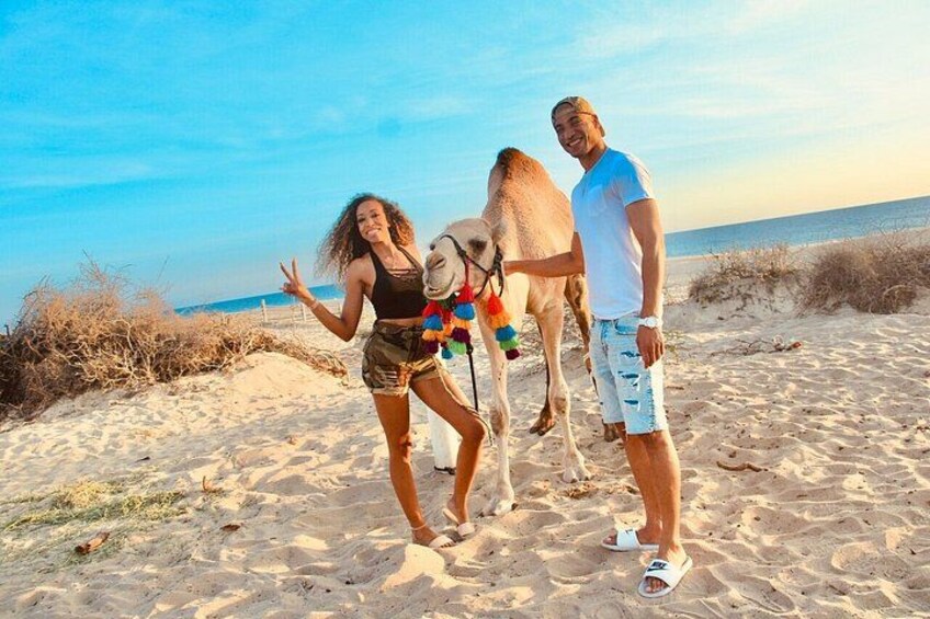 Una playa arenosa con un camello, una persona con camisa blanca y otra con remera negra. 3