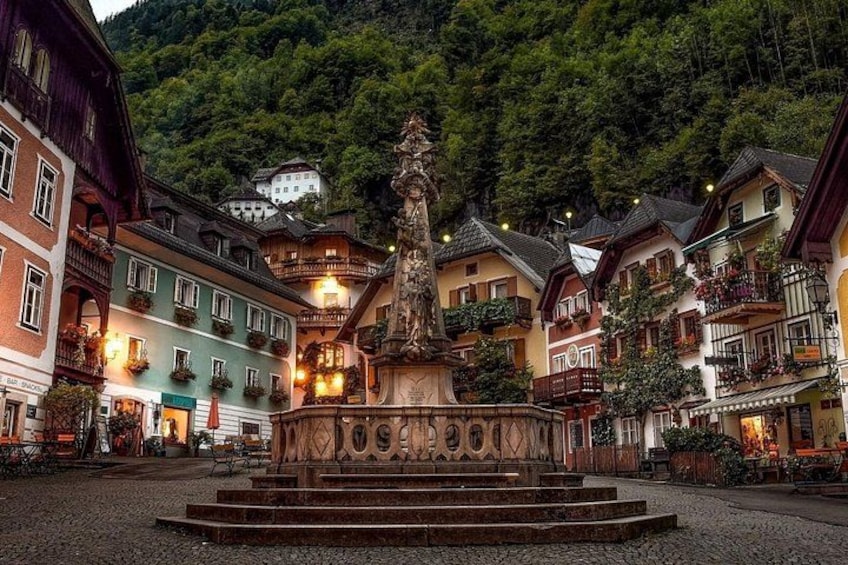 A town square with a central fountain, surrounded by colorful buildings and a forested hillside in the background. 3