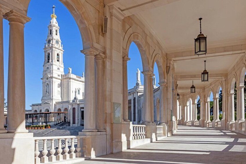 An architectural corridor with arches and columns, leading to a white church with a bell tower. 2