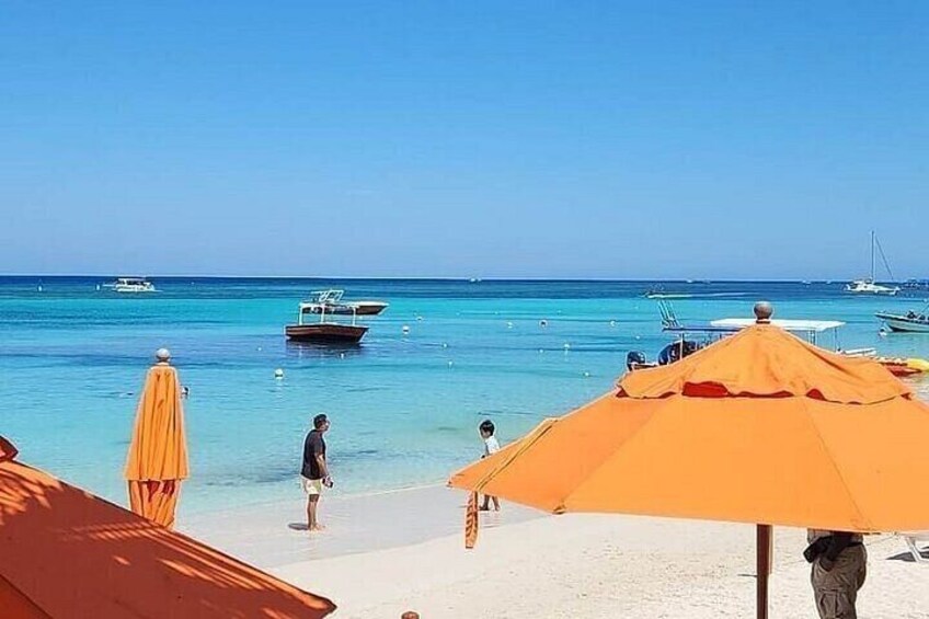 A beach with clear turquoise water, a few boats, and orange umbrellas. 2