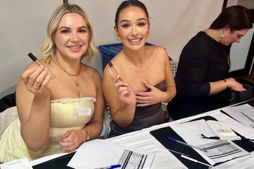 Two women holding makeup products at a table with papers and pens. 1