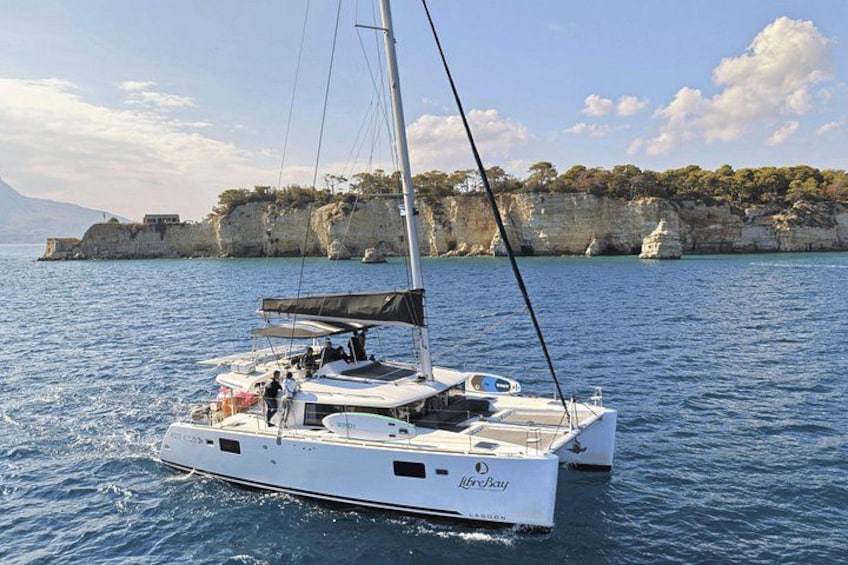 Un veliero bianco con cabina e albero, che naviga su acque blu calme con una costa rocciosa sullo sfondo. 1