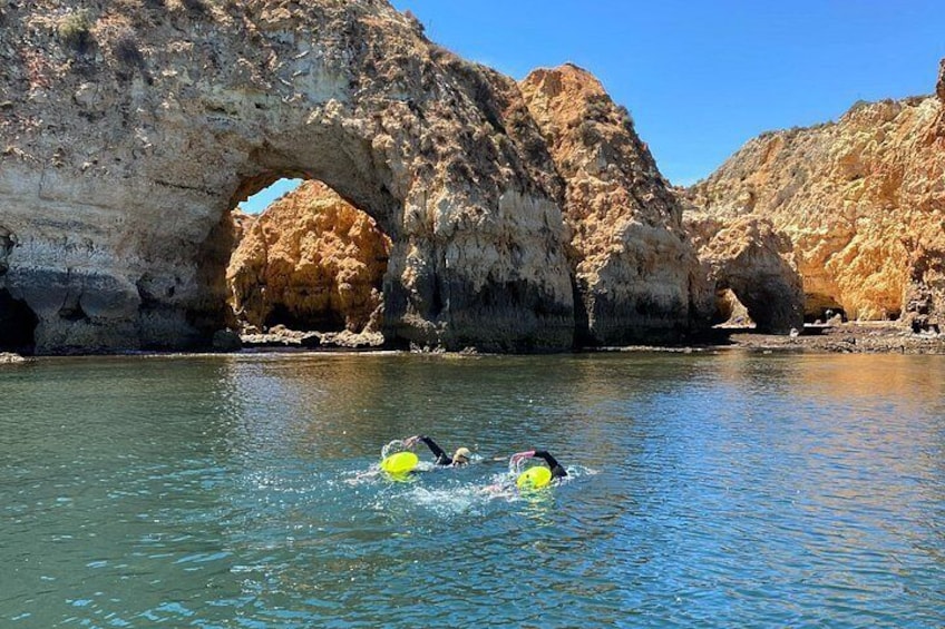 A natural rock formation with a large arch over water, two people swimming in the water, and a clear blue sky. 1