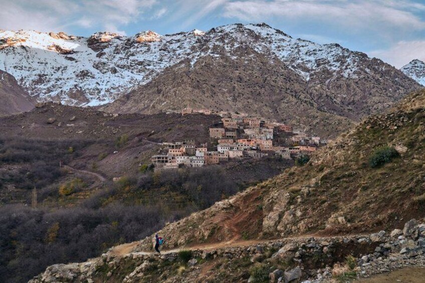 Ein Bergdorf mit Häusern, die sich im Tal befinden, eine Person wandert auf einem Pfad, und schneebedeckte Berge im Hintergrund. 5
