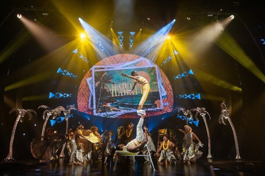 A stage with dancers in costumes, a large screen displaying 'Aloha Hawaii', and stage lighting. 5