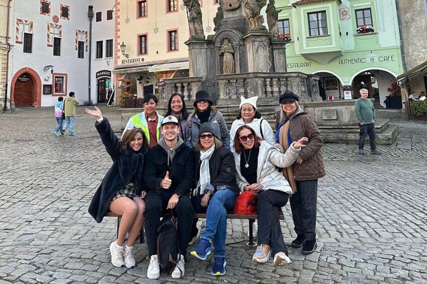 Un gruppo di persone che posano in una piazza lastricata, con edifici storici e una statua sullo sfondo. 2