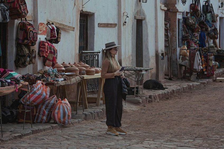A street market with goods displayed on tables, a person standing, and buildings with various items hanging on the walls. 1
