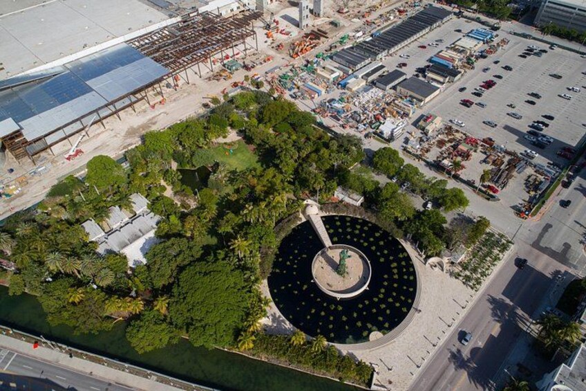 An aerial view of a park with a circular fountain, surrounded by trees and a parking lot. 3